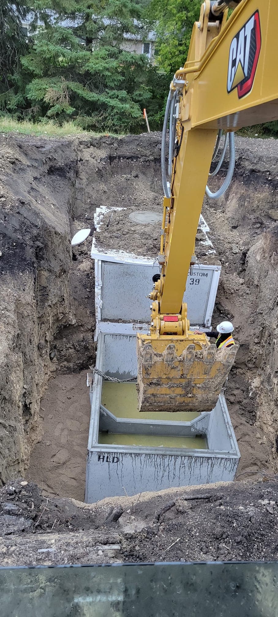 Yellow CAT excavator arm lowering concrete tanks into a deep trench at a construction site.