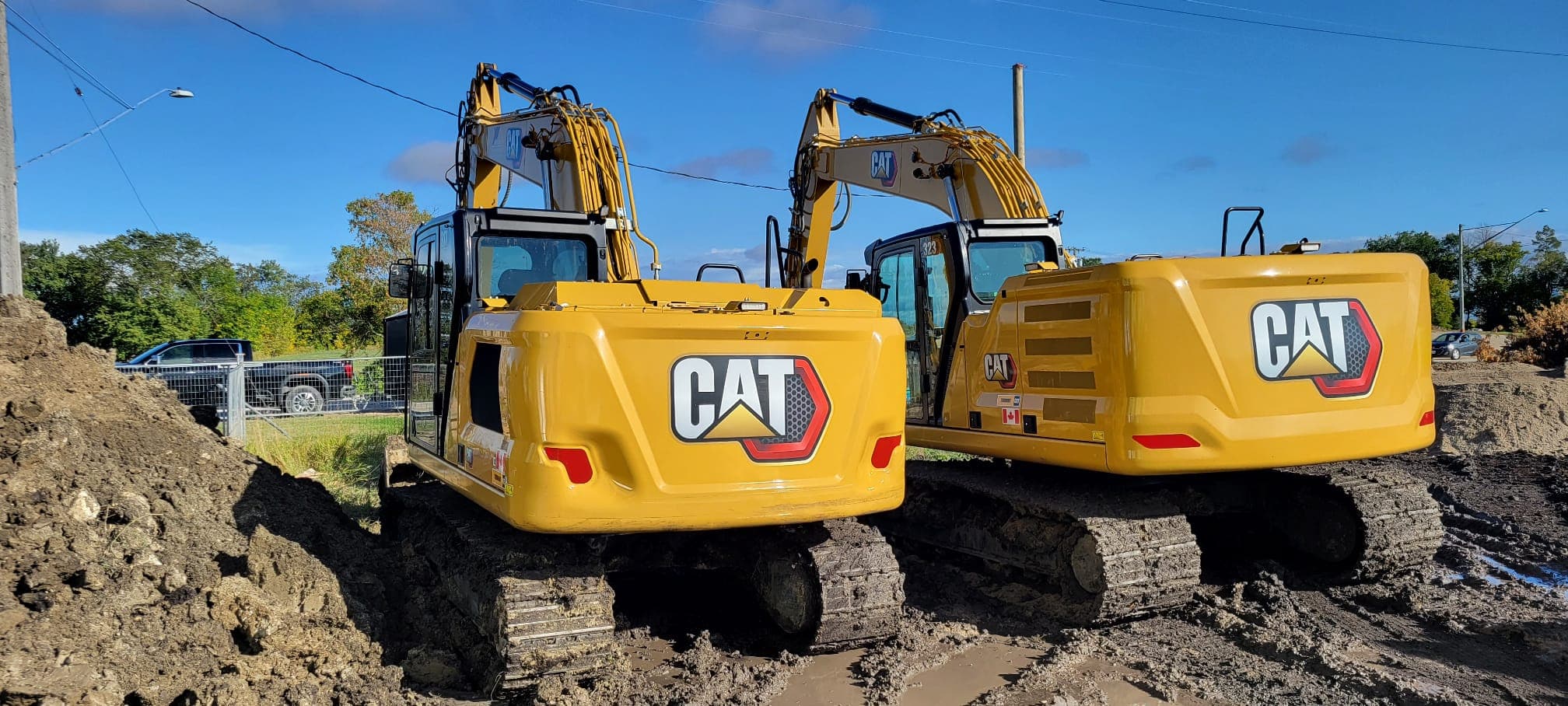 Rear view of two yellow Caterpillar excavators on a muddy construction site under blue skies.