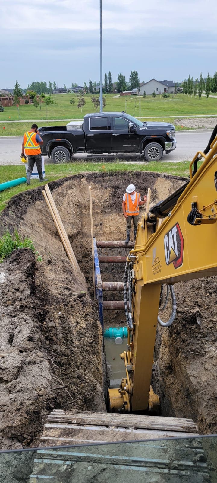 Yellow excavator arm lowers into a deep, shored trench with a worker standing inside.