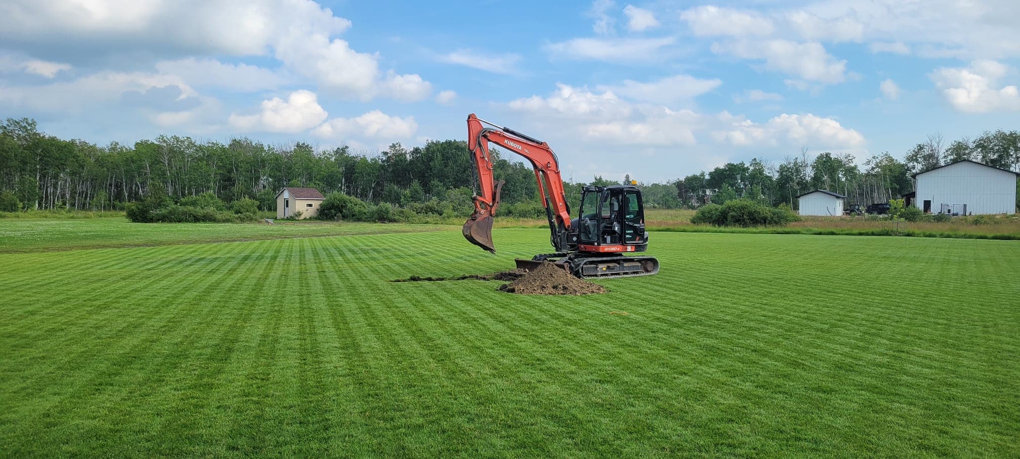 Orange Kubota excavator digging a hole in a striped green lawn with trees behind.