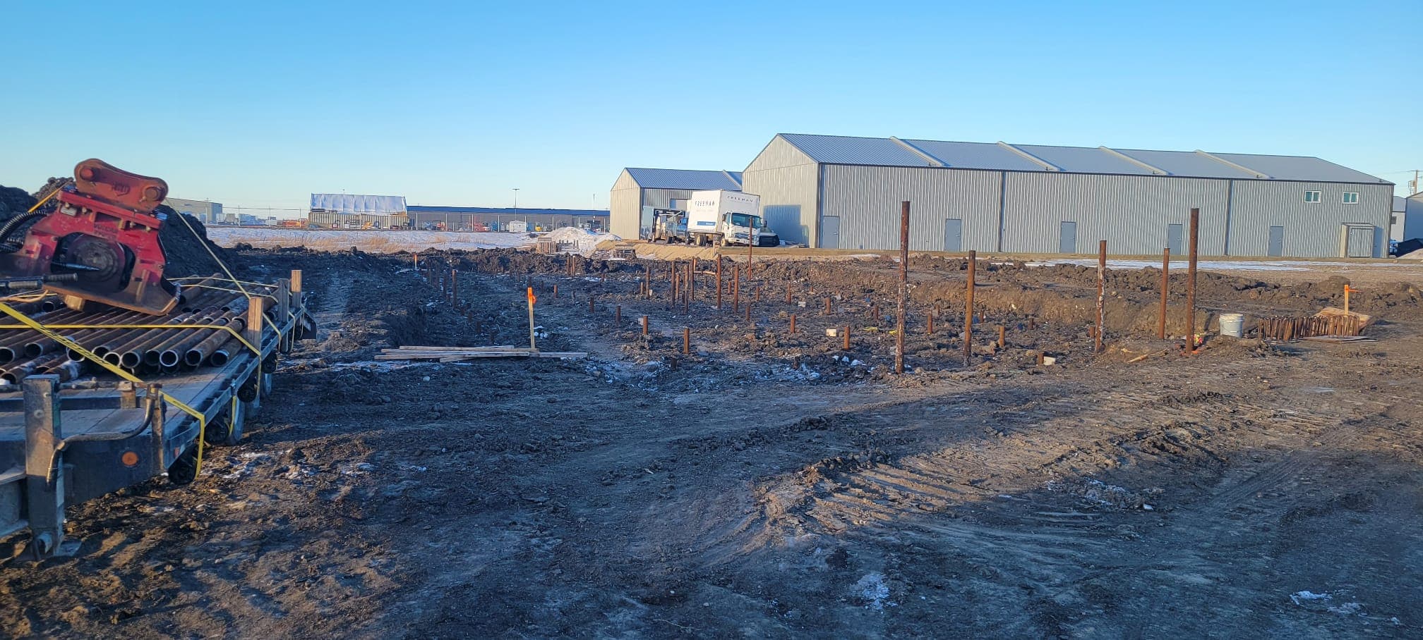 Vertical steel foundation piles driven into a muddy construction site near large industrial warehouse buildings.