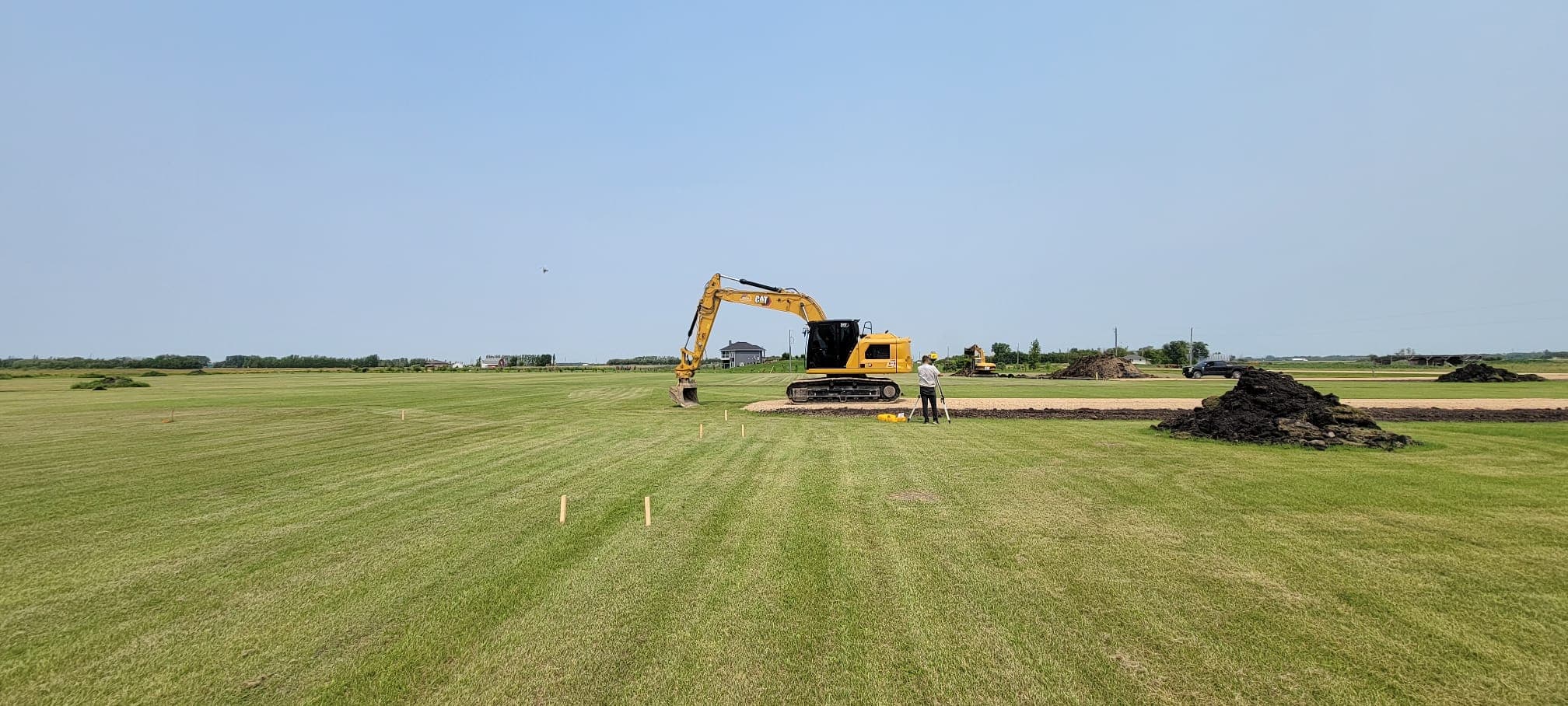 Yellow excavator and surveyor working in a vast green field under a clear sky.