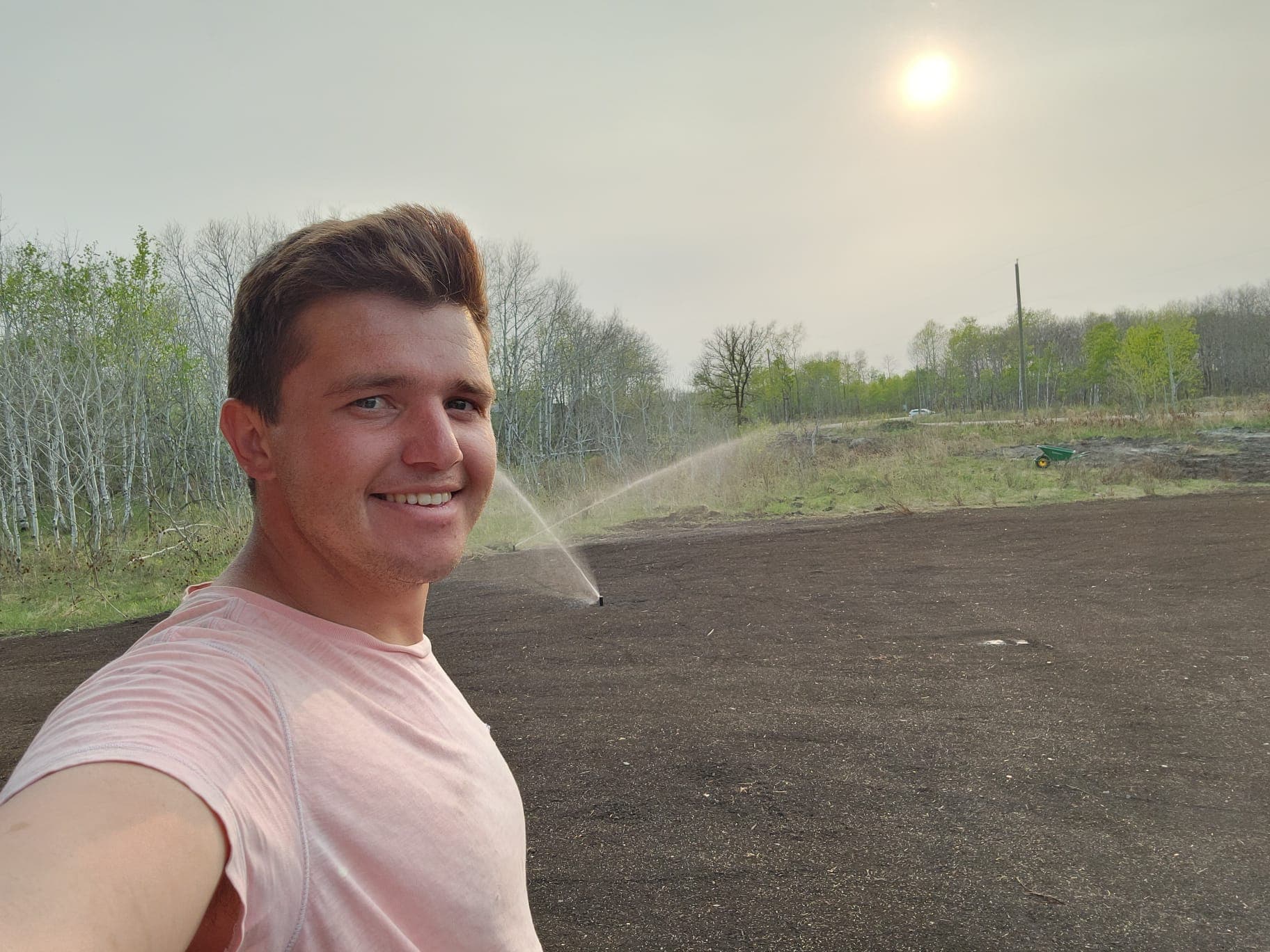 Smiling man takes a selfie beside a dirt field being watered by a sprinkler.