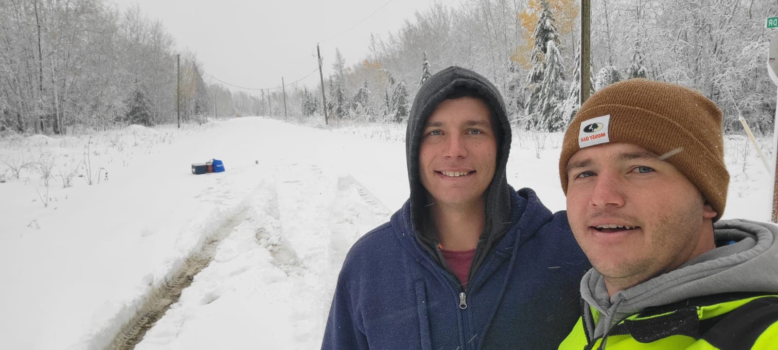 Two men smile while standing on a snow-covered road through a wintry forest.