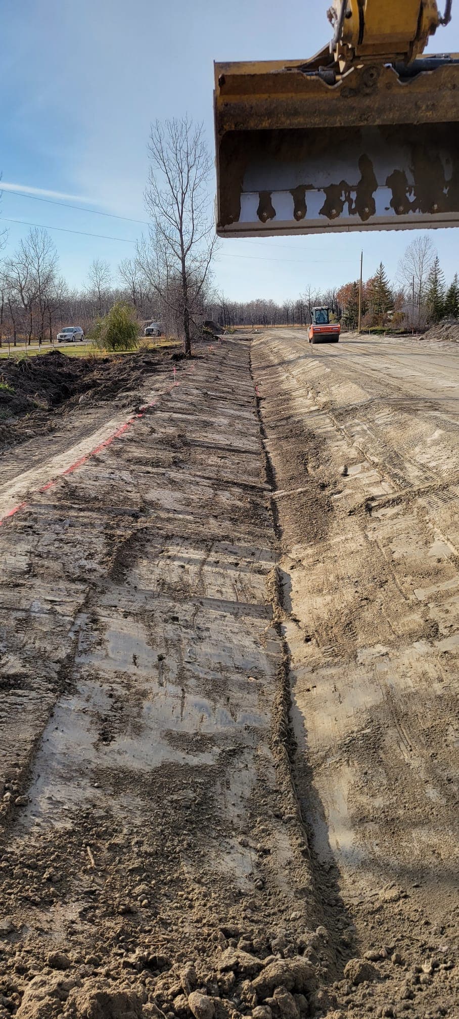 Graded dirt roadbed with a steamroller in the distance, viewed from an excavator bucket.